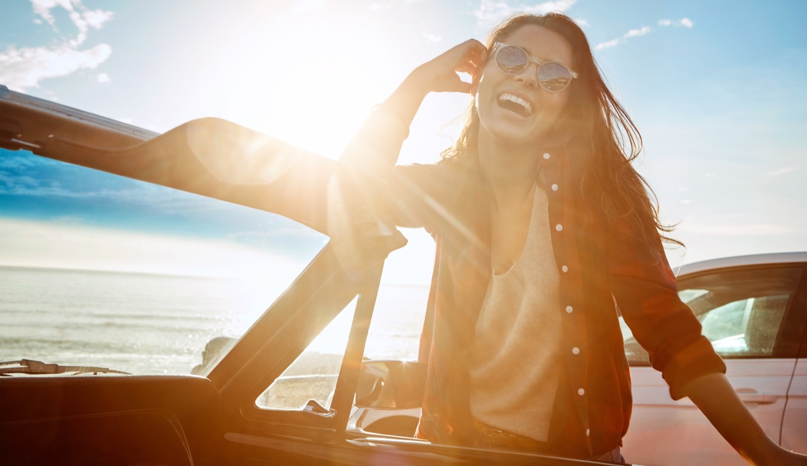 Woman laughing beside her car by the coast — car refinance eligibility